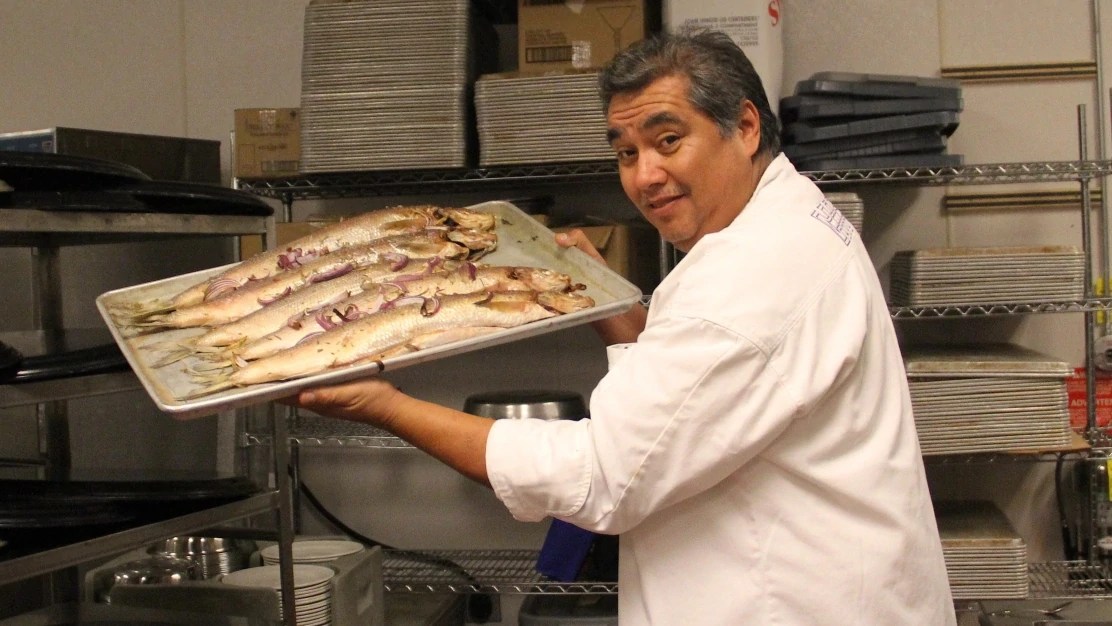 Chef Arlie Doxtator standing in a professional kitchen and holding a sheet pan with several whole fish on it