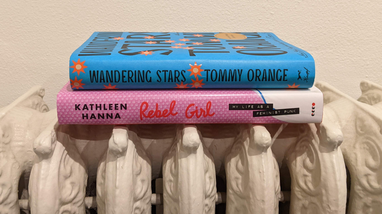 Two books stacked on a white radiator. The top book is Wandering Stars by Tommy Orange. The bottom book is Rebel Girl by Kathleen Hanna.